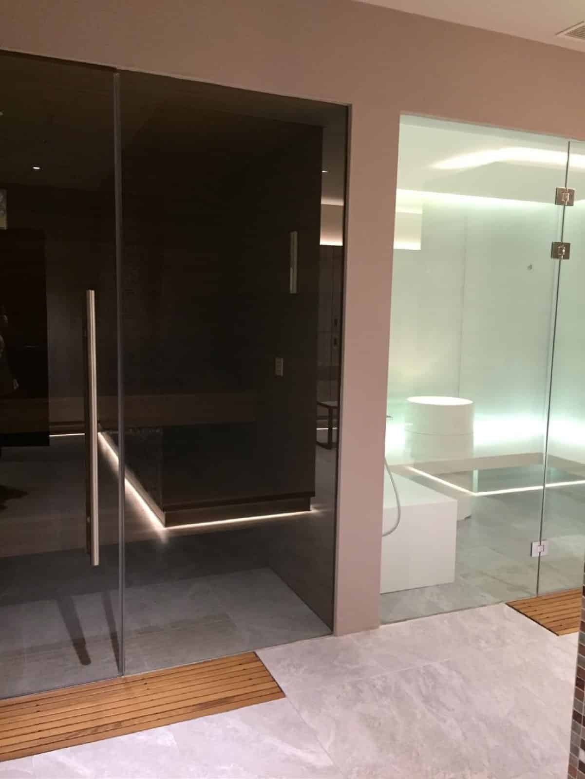 Wet-area corridor in an Emaar Square residence, Istanbul: the smoked glass front of a sauna with a long bar handle facing a white steam room behind clear glass, with timber duckboards on a pale stone floor