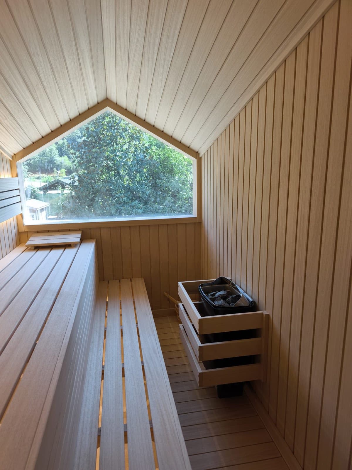 Wall-mounted stone sauna heater in a private villa sauna in Istanbul: stainless heater body filled with dark sauna stones, timber wall cladding, slatted bench above.