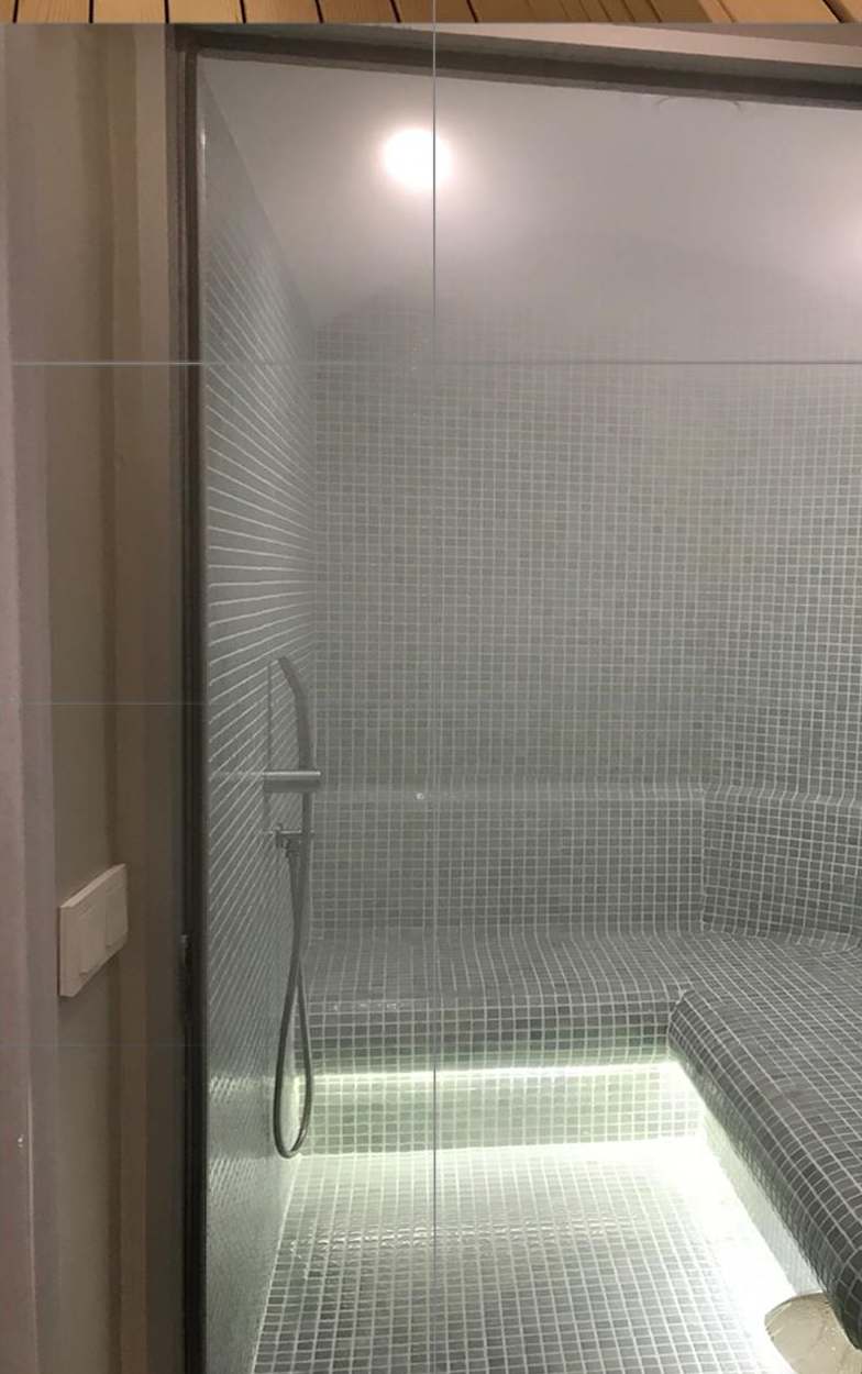Steam room at Oran Hotel, Istanbul, seen through its frameless glass front: pale grey mosaic walls, an L-shaped mosaic bench lit from below and a wall-mounted hand shower