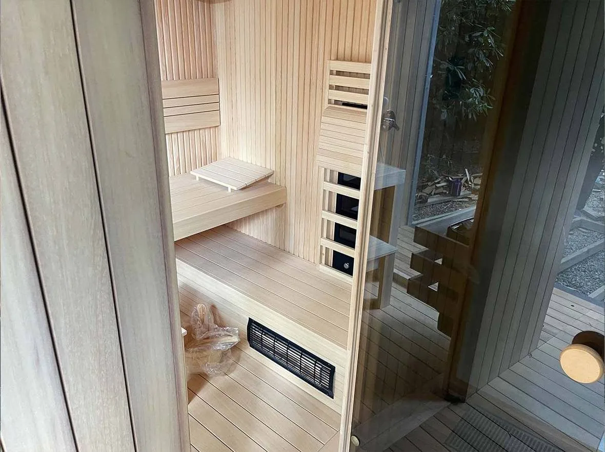 Small pale-timber sauna cabin interior: two bench tiers with a backrest, a glazed door, a low black exhaust grille in the bench riser, a control and speaker recess in the wall, timber decking and trees beyond the door