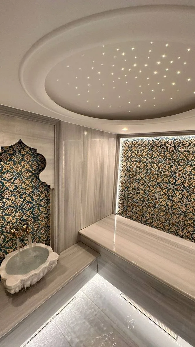 Corner of a hammam in grey figured marble with a white fluted kurna basin under a teal and gold tiled niche, a marble bench along the right wall, a domed ceiling recess with fibre optic star points and an illuminated linear drainage channel set into the floor