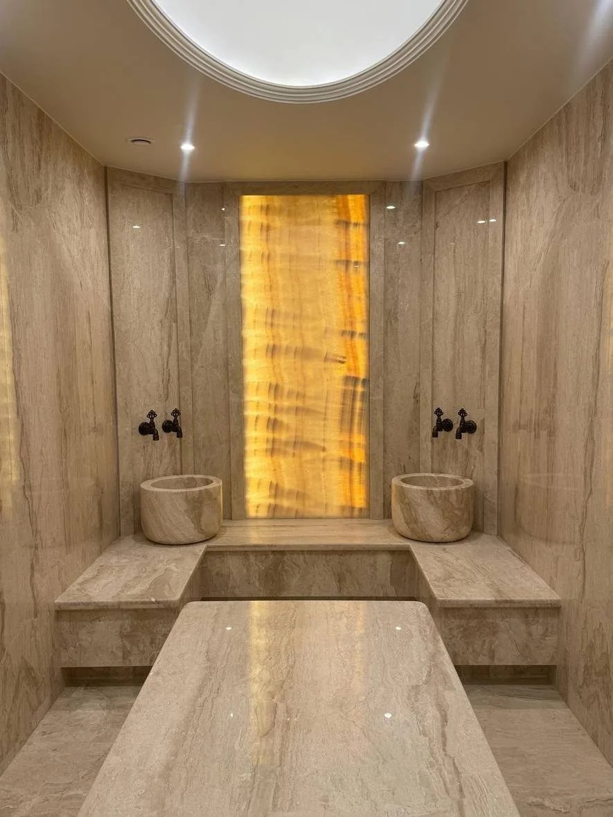 Private villa hammam in Nad Al Sheba, Dubai: veined pale stone walls, two stone basins with wall taps, a marble bench and a backlit amber onyx panel on the end wall