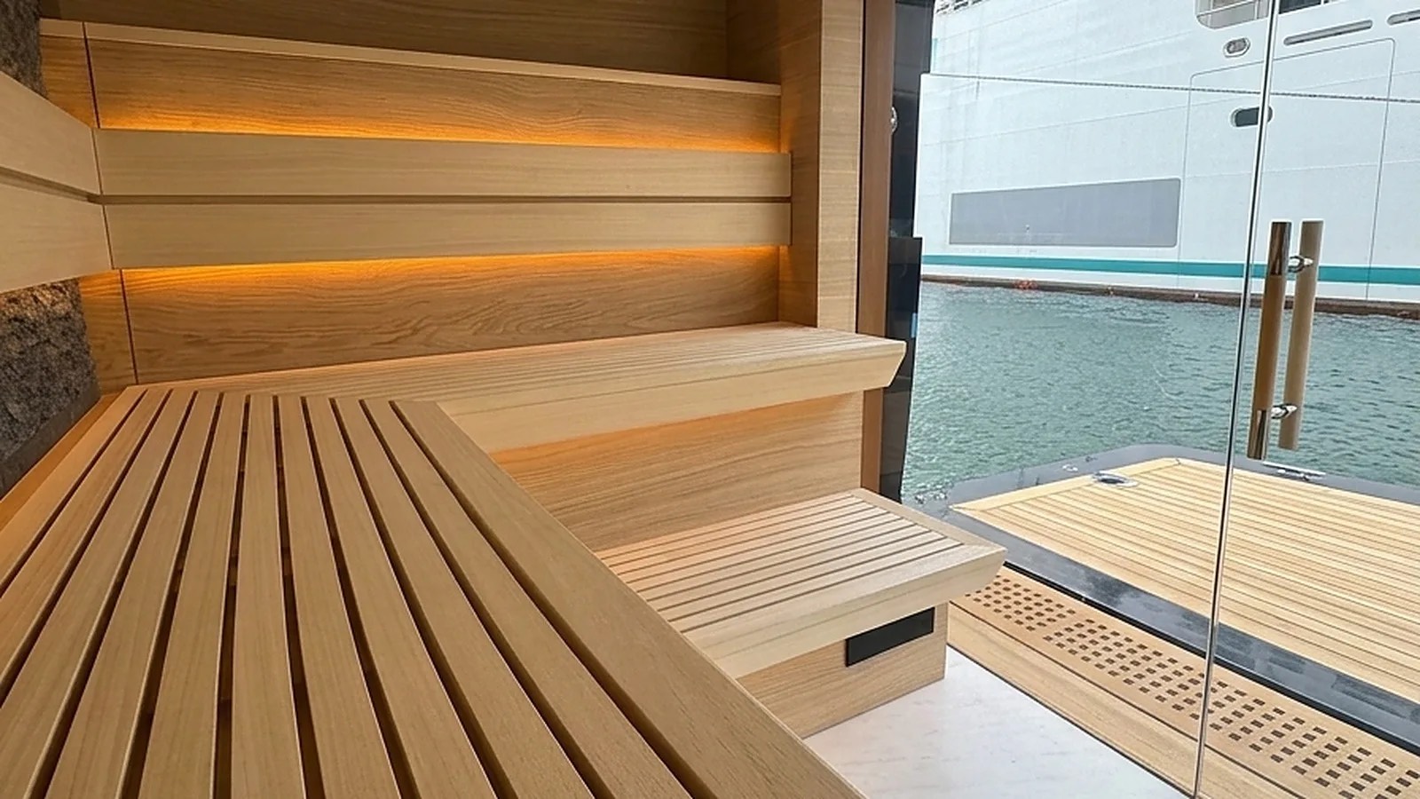 Sauna glass door and full-height clear glass front on a delivered cabin, pale timber benches with concealed strip lighting, looking out onto a marina
