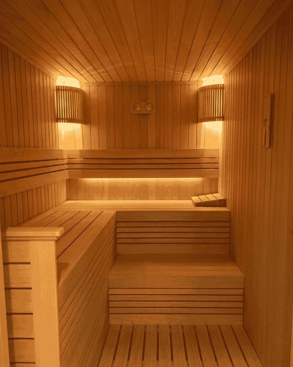 An L-shaped two-tier sauna room in pale timber with an upper bench, a lower step, two shaded wall lamps and a thermometer and hygrometer on the back wall
