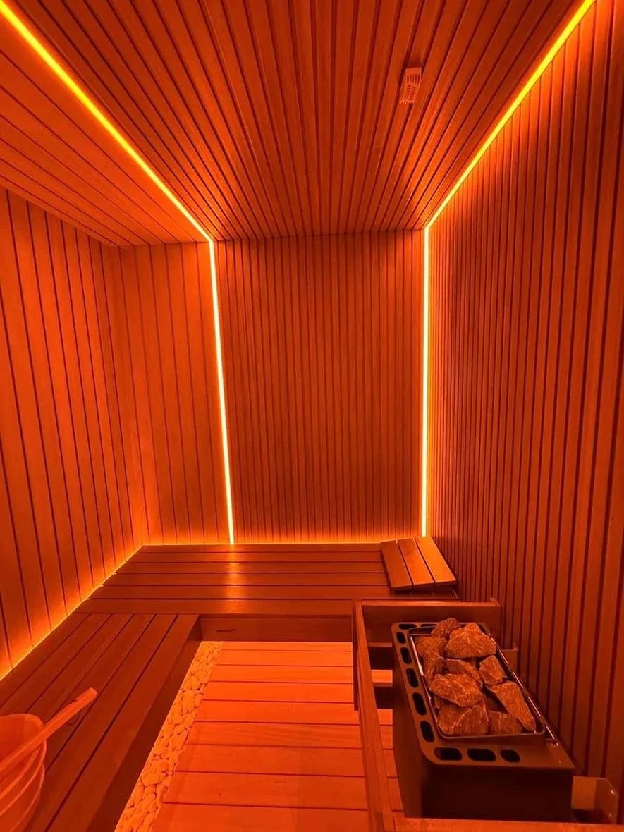 A timber sauna room seen from the door, with a light line running up each corner and along the ceiling, a low bench on two sides, a pebble strip on the floor and an electric heater behind a timber guard