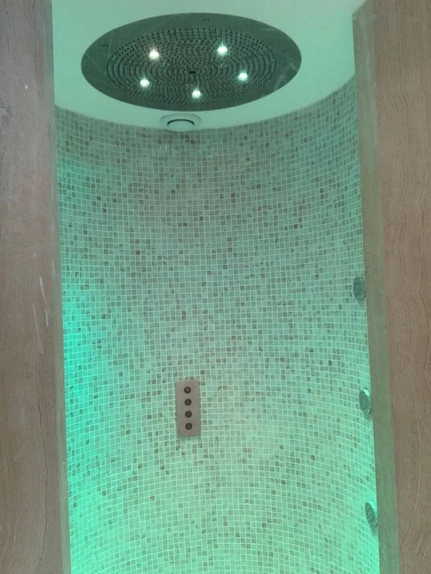 Round walk-in experience shower in a private villa on The Pearl, Doha, lined in pale mosaic with a round ceiling rain head, a wall control panel and green coloured lighting