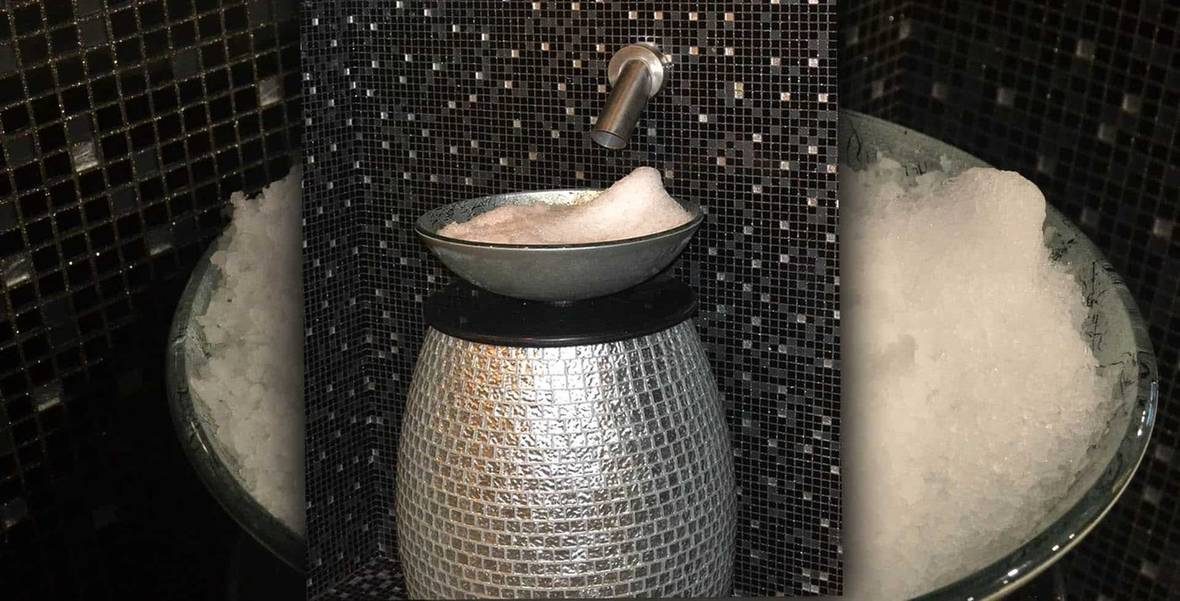 Ice fountain at Gumush Residence, Baku: a shallow bowl of flake ice on a silver mosaic pedestal under a stainless steel wall spout, set in a black mosaic niche