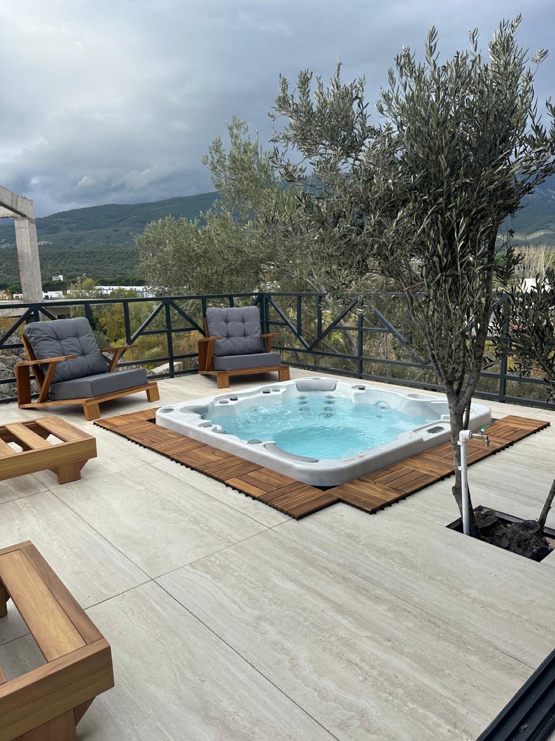 Rooftop terrace jacuzzi at a private forest retreat in the Kaz Mountains, Türkiye: a white acrylic tub set into pale stone paving, framed with timber deck tiles, beside an olive tree in a planter and a metal railing